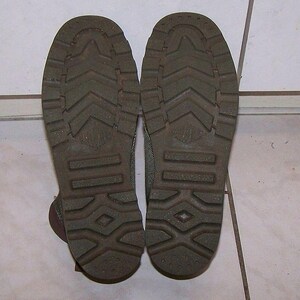 May include: Close-up of the soles of two olive green boots. The soles have a textured pattern with a diamond and arrow design. The boots are placed on a white tiled surface.