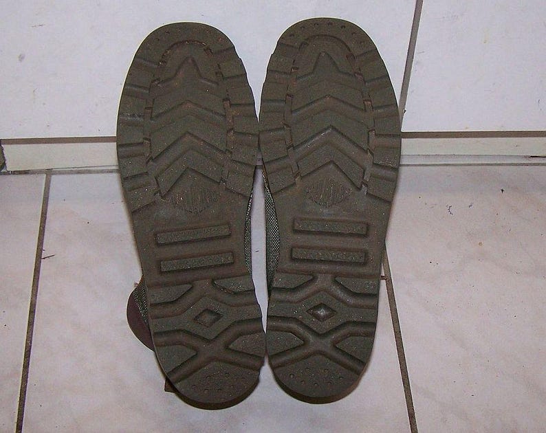 May include: Close-up of the soles of two olive green boots. The soles have a textured pattern with a diamond and arrow design. The boots are placed on a white tiled surface.
