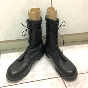 Puede incluir: Un par de botas de combate de cuero negro con cordones, con punta redondeada y suela gruesa. Las botas están en posición vertical, con hormas beige en el interior. Las botas están sobre un suelo embaldosado.