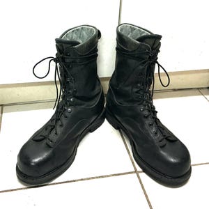 May include: A pair of black leather and fabric tactical boots featuring a high-top design, black laces, metal eyelets, and a light grey quilted interior lining. The boots have a rounded toe, a thick black rubber sole, and a sturdy construction.