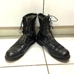 Könnte beinhalten: Paar schwarze Leder-Kampfstiefel mit Frontreißverschlüssen und Schnürungen. Die Stiefel haben ein abgenutztes Aussehen, was darauf hindeutet, dass sie Vintage sind oder benutzt wurden. Die Sohlen sind dunkel und die Stiefel stehen auf einer weißen Fliesenoberfläche.