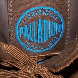 May include: Close-up of a brown fabric patch with the blue text "PALLADIUM" inside a blue oval. The text "L'ORIGINALE DEPUIS 1947" is arched around the oval. Brown shoelaces are visible in the foreground.