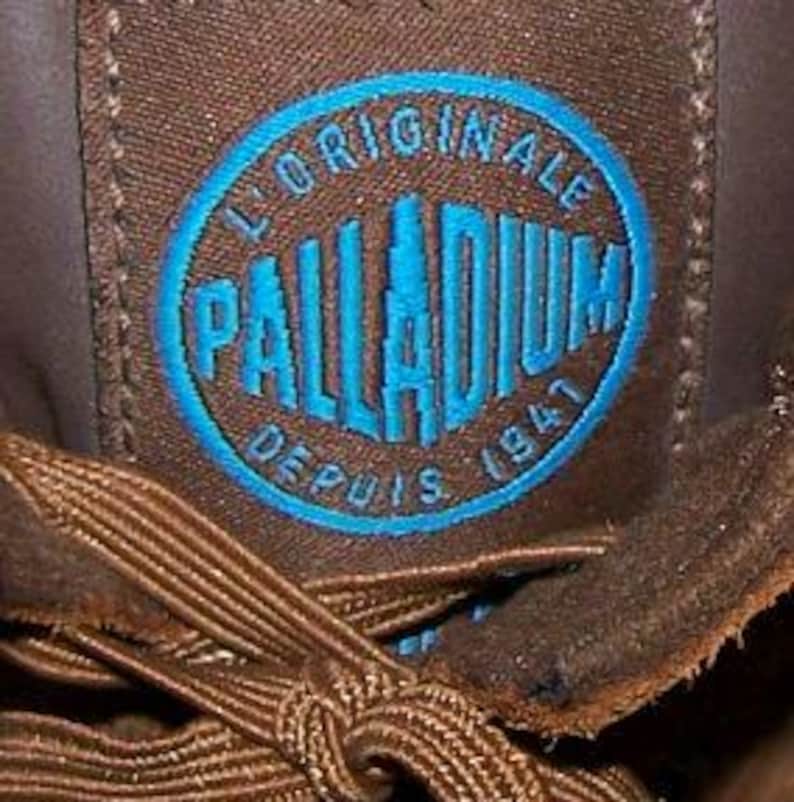 May include: Close-up of a brown fabric patch with the blue text "PALLADIUM" inside a blue oval. The text "L'ORIGINALE DEPUIS 1947" is arched around the oval. Brown shoelaces are visible in the foreground.
