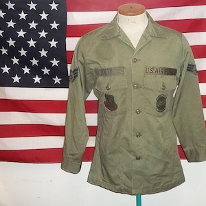 Könnte beinhalten: Ein olivgrünes Uniformhemd der US Air Force mit den Aufnähern "SOETY" und "U.S. AIR FORCE". Das Hemd hat zwei Brusttaschen und wird vor einer amerikanischen Flagge präsentiert.