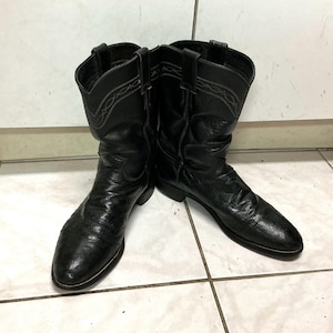 Op de afbeelding: Een paar zwarte leren cowboylaarzen met een textuurafwerking, ronde neuzen en treklussen aan de bovenkant van de schachten. De bovendelen van de laarzen zijn versierd met subtiele, lichtgekleurde stikpatronen langs de rand.