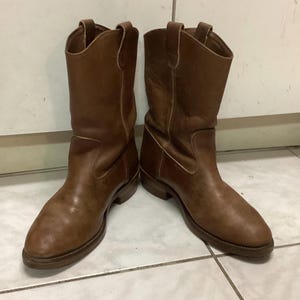 Puede incluir: Un par de botas de cuero marrón a media pantorrilla con punta redondeada, tacón bajo y tiradores en la parte superior de la caña. Las botas tienen costuras visibles a los lados y un acabado mate liso en toda la superficie exterior.
