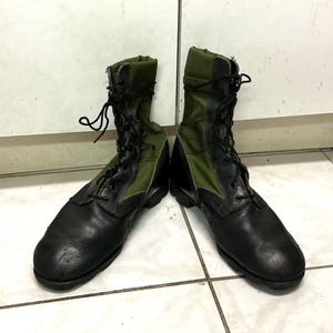 May include: A pair of high-top combat boots featuring black leather toe caps and heel counters, combined with olive green canvas side panels. The boots include black laces, metal eyelets, and a thick black rubber sole for traction and durability.
