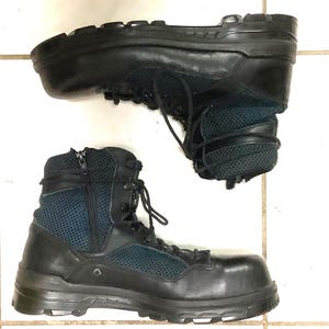 May include: A pair of black tactical boots featuring a combination of smooth leather and dark blue mesh panels. These high-top boots include a side zip closure, black laces, metal eyelets, and a thick, lugged rubber sole for traction.