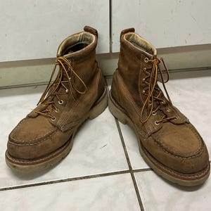 May include: A pair of brown leather work boots featuring a moc-toe design, high-top ankle support, and thick, light-coloured rubber soles. The boots are secured with yellow and brown braided laces threaded through metal eyelets and hooks.
