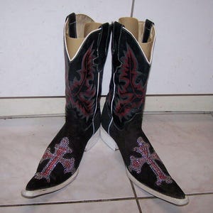 May include: A pair of black cowboy boots with a pointed toe and a white sole. The boots feature a red and grey floral design on the shaft and a cross design on the toe, embellished with rhinestones.