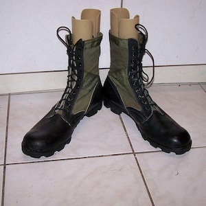 May include: A pair of black and olive green combat boots with black laces. The boots have a rubber sole and canvas and leather uppers. Boot trees are inserted in the boots.