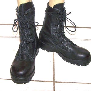 Puede incluir: Un par de botas de combate de cuero negro con diseño de caña alta, punteras redondeadas y suelas gruesas de goma con tacos. Las botas se aseguran con cordones negros que pasan por ojales metálicos y ganchos hasta la media pantorrilla.