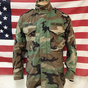 May include: Camouflage military jacket with the name "WEINSTEIN" and "U.S. ARMY" patches. The jacket features a high collar, multiple pockets, and a button-up front. The background is an American flag.