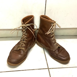 Könnte beinhalten: Ein Paar braune Lederstiefel mit hellen Schnürsenkeln. Die Stiefel haben eine Mokassin-Zehenform und eine mittlere Wadenhöhe. Die Sohlen sind hellbraun. Die Stiefel sind geschnürt und scheinen in gutem Zustand zu sein.