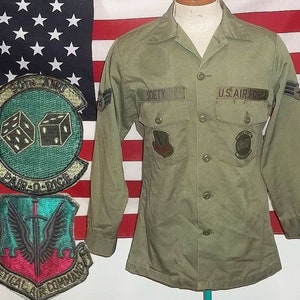 Puede incluir: Una camisa de uniforme verde oliva de la Fuerza Aérea de los EE. UU. con el nombre "SOETY" encima del bolsillo izquierdo. Los parches incluyen "PAIR-O-DICE" y "TACTICAL AIR COMMAND". La bandera estadounidense está al fondo.