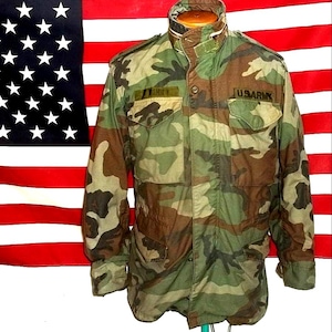 May include: A camouflage field jacket featuring a high collar, long sleeves, and two chest pockets. The jacket displays a woodland pattern in shades of green, brown, and black. A patch reading U.S. ARMY is visible on the right chest area.
