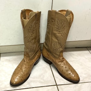 May include: A pair of tan cowboy boots featuring a textured, quill-patterned leather vamp and a smooth leather shaft with decorative stitching. The boots have a pointed toe, a stacked heel, and pull tabs at the top of the tall, structured shafts.