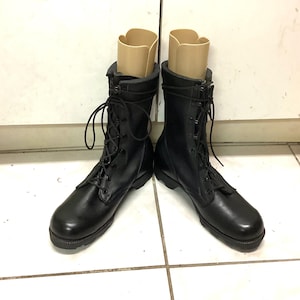 Puede incluir: Un par de botas de combate de cuero negro con diseño de cordones. Las botas tienen una puntera redondeada y una suela resistente. Se insertan hormas beige dentro de las botas. Las botas están sobre un suelo de baldosas blancas.