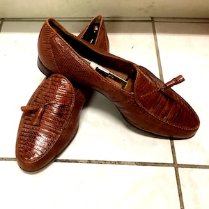 Pode incluir: Um par de mocassins castanhos com um padrão texturizado de pele de lagarto e borlas decorativas no peito do pé. Os sapatos são construídos com um salto baixo e uma biqueira arredondada, exibindo um acabamento brilhante e silhueta fina.
