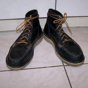 May include: A pair of black leather work boots with tan soles and yellow and brown laces. The boots have a moccasin-style toe and are laced up. The boots are sitting on a tiled surface.