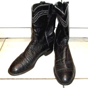Könnte beinhalten: Ein Paar schwarze Cowboy-Stiefel mit strukturiertem Lederfinish. Die Stiefel haben eine spitze Zehenpartie und ein dekoratives weißes Nahtmuster am oberen Rand. Die Stiefel sind aus Leder.