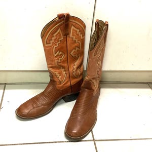 Op de afbeelding: Een paar bruine leren cowboylaarzen met een textuur op de wreef en een lichtere beige schacht met decoratieve zigzagstiksels. De laarzen hebben een klassieke spitse neus, een hak en treklussen aan de bovenkant van de schacht.