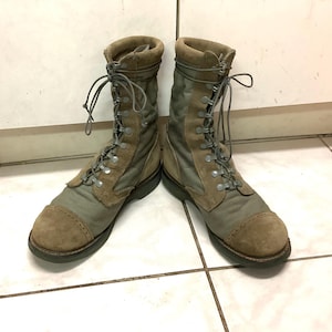 May include: A pair of olive green and tan tactical boots featuring a high-top design with lace-up closures, reinforced suede toe caps, and durable fabric side panels. The boots have metal eyelets and sturdy rubber soles, providing a structured fit.