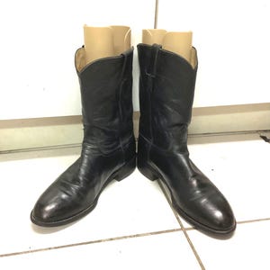 Op de afbeelding: Een paar zwarte leren cowboylaarzen. De laarzen hebben een klassiek ontwerp met spitse tenen en een middelhoge kuit. Het leer lijkt gepolijst en de laarzen zijn in goede staat. De laarzen staan rechtop op een wit betegeld oppervlak.