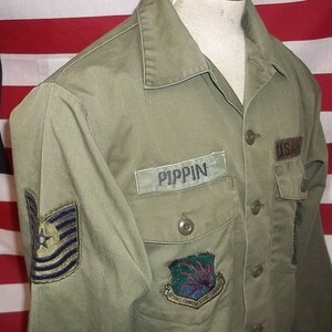 Puede incluir: Una camisa de uniforme militar verde oliva con el nombre "PIPPIN" en una etiqueta con el nombre. La camisa tiene dos bolsillos en el pecho, un parche "USAF" y un parche colorido con una estrella y rayas. La camisa está abotonada y expuesta en un maniquí.