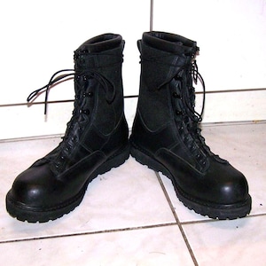 May include: A pair of black tactical boots featuring a smooth leather toe cap, a high-top shaft with fabric side panels, black laces threaded through metal eyelets, and a thick, lugged rubber outsole for traction and stability.