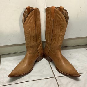May include: A pair of light brown cowboy boots. The boots have pointed toes, decorative stitching, and pull straps at the top. The leather boots are standing on a tiled surface.