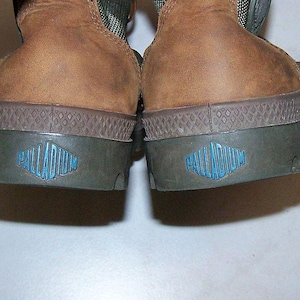May include: Close-up of the heels of two brown leather boots. The soles are dark green with a diamond pattern and a blue logo that reads "PALLADIUM". The boots have a rugged, worn appearance.