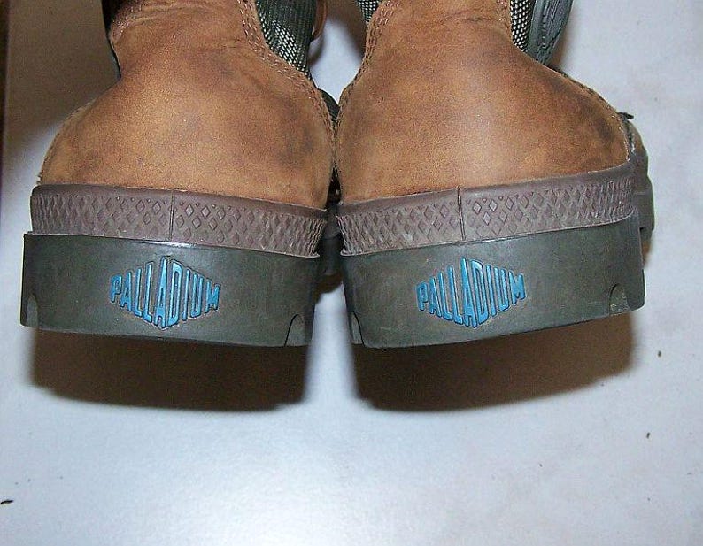 May include: Close-up of the heels of two brown leather boots. The soles are dark green with a diamond pattern and a blue logo that reads "PALLADIUM". The boots have a rugged, worn appearance.