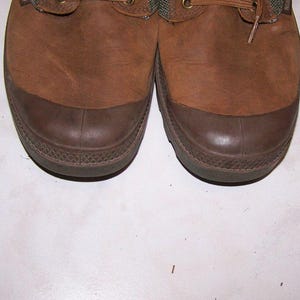 May include: Pair of brown leather boots with dark brown rubber toe caps. The boots have a textured appearance and are laced up with brown laces. The soles are a darker shade of brown.