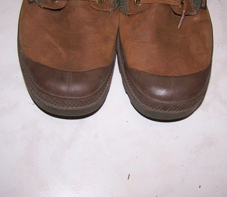 May include: Pair of brown leather boots with dark brown rubber toe caps. The boots have a textured appearance and are laced up with brown laces. The soles are a darker shade of brown.