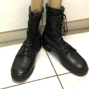 Könnte beinhalten: Ein Paar schwarze Leder-Kampfstiefel mit hohem Schaft, abgerundeten Zehenkappen und schwarzen Schnürsenkeln, die durch Metallösen geführt werden. Die Stiefel haben eine robuste Konstruktion mit einer dicken Sohle und einer glatten Oberfläche.