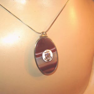 Pode incluir: Colar com pingente oval e corrente prateada. O pingente apresenta uma pedra polida, listrada em bordô e branco, com uma moldura prateada. Um pequeno emblema circular com um navio e uma âncora está centrado na pedra.