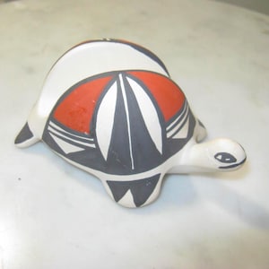 May include: A ceramic turtle figurine featuring a smooth white base with hand-painted geometric patterns in matte black and vibrant orange. The shell displays symmetrical triangular and linear motifs, while the head has simple black eye markings.