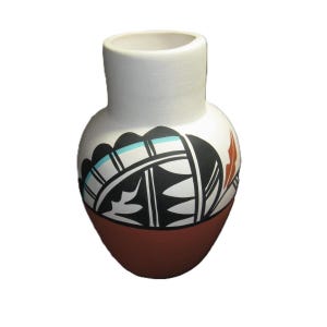 Puede incluir: Un pequeño jarrón de cerámica hecho a mano con una sección superior blanca y una base marrón rojizo. El jarrón presenta un diseño geométrico en blanco y negro con detalles turquesa y naranja, mostrando una estética tradicional.