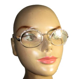 May include: Silver-rimmed, round eyeglasses with clear lenses. The frames have a decorative, beaded design along the top edges. The glasses are displayed on a mannequin head with a neutral skin tone.