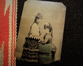 May include: An antique sepia-toned photograph of two women in Victorian-era clothing. The woman in the front is wearing a dark dress with a white lace trim and a hat. The woman in the back is wearing a light-colored dress with a dark trim and a hat.