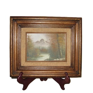 May include: A framed landscape painting depicting a mountain scene with a river and trees. The painting is in a wooden frame with a burlap-like border. The frame is displayed on a dark brown easel.