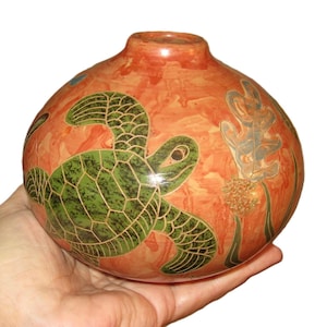 May include: A round, orange ceramic vase with a green sea turtle design. The turtle is detailed with a dark green pattern. The vase also features a design of seaweed and other aquatic plants.