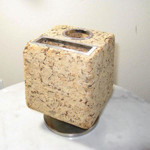 May include: A square-shaped desk organizer made of textured light brown cork. The top surface features a rectangular metal slot and a circular metal opening. The entire structure is supported by a circular, polished silver-toned metal base.