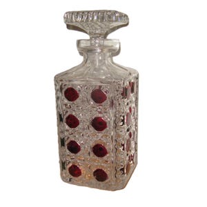 May include: A clear glass decanter with a square body and a decorative stopper. The decanter features a pattern of red circular accents. The stopper is also clear and has a textured design. The overall design is vintage.