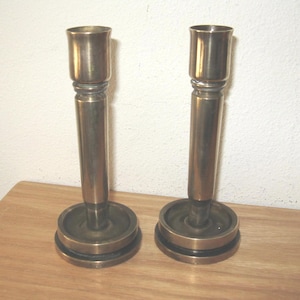 May include: Two antique brass candlesticks. Each candlestick has a round base, a long cylindrical stem, and a cup-shaped top to hold a candle. The candlesticks are made of a shiny, golden-colored metal and are displayed on a wooden surface.