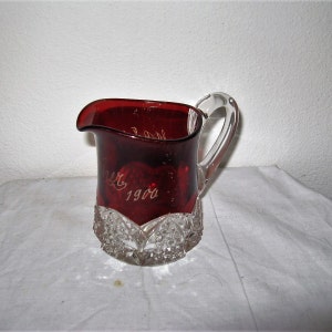 Puede incluir: Una jarra de vidrio roja con un asa de vidrio transparente. La jarra tiene una base de vidrio transparente con un diseño de vidrio cortado. La jarra tiene las palabras "Año 1900" grabadas en el lateral.