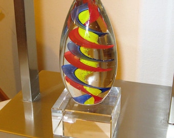 May include: A glass paperweight with a swirling pattern of red, blue, and yellow. The paperweight is on a clear glass base.
