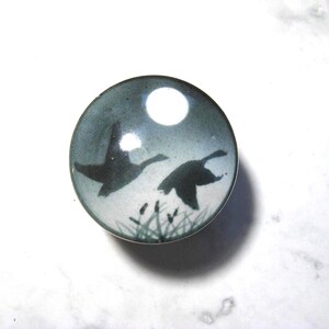 May include: A circular cabinet knob with a smooth, glossy finish featuring a printed graphic of two dark silhouettes of birds in flight against a pale blue and white background, with a small cluster of dark reeds depicted at the bottom edge.