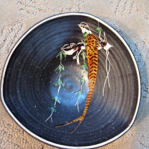 Könnte beinhalten: Eine schwarze Keramikschale mit weißem Rand zeigt ein handgemaltes Design eines Fasanen mit langem Schwanz und grünen Blättern. Die Schale hat eine gesprenkelte schwarze Glasur.